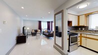 This well-appointed single-floor residence offers 2 bedrooms and 1.5 bathrooms, thoughtfully designed with a modern aesthetic and functional open-concept layout. The living area is expansive and welcoming, featuring gleaming large-format white tile flooring that reflects natural light throughout the space. Recessed ceiling lighting provides bright, even illumination, complementing the natural light that filters in through windows dressed with rich, deep-purple curtains that add a bold accent to the neutral palette. The living room comfortably accommodates dual seating areas, creating a versatile space for both relaxing and entertaining. A full-length mirror with a rustic wood frame serves as a stylish architectural accent, visually expanding the space further. Adjacent to the living area, the kitchen is efficiently designed and features stainless steel appliances, including a range and microwave, white cabinetry, granite-style countertops, and a mosaic tile backsplash that adds texture and character. The seamless flow between the kitchen and living spaces makes this home ideal for everyday living and hosting. The overall ambiance is clean, contemporary, and bright, with a cohesive design language carried throughout the single-level floor plan.