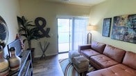 This single-floor space offers a comfortable and well-appointed living environment designed with both style and functionality in mind. The living area serves as the heart of the space, showcasing rich hardwood-style flooring that adds warmth and character throughout. A large sliding glass door floods the room with natural light and provides direct access to an outdoor balcony, offering views of the surrounding exterior. Vertical blinds along the sliding door allow for flexible light control and privacy. The living area is generously sized, accommodating a tufted leather sofa, ottomans, and a decorative area rug with multicolored striped accents. Neutral-toned walls complement the earthy furnishings and decorative elements, including a prominent wall-mounted ampersand symbol and a tall indoor plant that adds a touch of greenery. A floor lamp in the corner provides additional ambient lighting, enhancing the cozy atmosphere of the room. The overall design aesthetic is casual yet put-together, making the space feel welcoming and livable. The seamless connection between the interior living area and the outdoor balcony is a standout feature, contributing to an open, airy feel throughout.