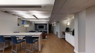 This single-floor space offers a well-appointed interior that blends functional work areas with comfortable lounge settings. The layout is open and expansive, featuring two distinct ceiling styles: an exposed beam section with industrial-style linear lighting over the main area, and an ornate tin ceiling detail near the reception or workstation zone, adding architectural character to the space.

A large stone-topped counter with bar-height seating serves as a central gathering or collaborative work surface, complemented by a dedicated workstation area along one wall, complete with cabinetry, a computer setup, and a coffee station. The warm hardwood flooring flows throughout, grounding the space with a welcoming, cohesive aesthetic.

Toward the rear, a cozy lounge area is defined by a blue accent wall, wall-mounted television, and comfortable seating, creating a relaxed setting for informal meetings or downtime. Soft wall sconces and a floor lamp provide warm ambient lighting throughout. The painted brick walls and neutral gray tones contribute to a modern yet inviting atmosphere, making this a versatile and well-designed space for both productivity and relaxation.