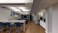 This single-floor space offers a well-appointed interior that blends functional work areas with comfortable lounge settings. The layout is open and expansive, featuring two distinct ceiling styles: an exposed beam section with industrial-style linear lighting over the main area, and an ornate tin ceiling detail near the reception or workstation zone, adding architectural character to the space.

A large stone-topped counter with bar-height seating serves as a central gathering or collaborative work surface, complemented by a dedicated workstation area along one wall, complete with cabinetry, a computer setup, and a coffee station. The warm hardwood flooring flows throughout, grounding the space with a welcoming, cohesive aesthetic.

Toward the rear, a cozy lounge area is defined by a blue accent wall, wall-mounted television, and comfortable seating, creating a relaxed setting for informal meetings or downtime. Soft wall sconces and a floor lamp provide warm ambient lighting throughout. The painted brick walls and neutral gray tones contribute to a modern yet inviting atmosphere, making this a versatile and well-designed space for both productivity and relaxation.
