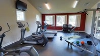 This single-story fitness center offers a comprehensive workout environment designed to accommodate a variety of exercise routines. The space is defined by vaulted ceilings that enhance the sense of openness, while bold red accent walls provide an energizing atmosphere against the neutral cream-colored upper walls. Multiple windows along the far wall flood the room with natural light and offer views of the surrounding outdoor greenery, creating an inviting and motivating environment.

The cardio zone is well-stocked, featuring elliptical trainers and treadmills arranged to maximize floor space and allow comfortable movement between machines. Protective floor mats are positioned beneath each piece of equipment. Along the far wall, a dedicated free weights area includes a full dumbbell rack with varying weights alongside a weight bench, catering to strength training needs. The open floor area provides ample room for stretching and functional training exercises, with a yoga mat, medicine ball, and additional accessories available for use. A wall-mounted television is positioned for easy viewing during workouts. Overhead fluorescent lighting supplements the natural light, ensuring the space remains bright throughout the day.