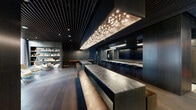 Located at 45 Lansing St in San Francisco's vibrant SoMa district, this meticulously designed single-floor space offers a generously proportioned interior that blends modern sophistication with refined materiality. The open-plan layout is anchored by striking dark wood paneling, a slatted ceiling detail, and rich hardwood flooring that runs throughout, creating a cohesive and moody aesthetic.

A central gathering area features elongated marble-topped communal tables paired with bench seating, positioned beneath a sculptural branching light installation that serves as a dramatic focal point. Adjacent to this is a full bar counter lined with sleek black stools, backed by a large mirror and integrated display screens — ideal for events and social gatherings.

A dedicated catering or prep kitchen features dual runs of veined marble countertops with undermount sinks, dark cabinetry, and pendant lighting, offering both functionality and visual continuity with the main space.

Toward one end, a bright lounge area with floor-to-ceiling windows admits abundant natural light, complemented by modern swivel chairs and built-in shelving. Glass partitions with gold-toned frames define zones while maintaining visual openness, and elegant drapery adds softness throughout. The overall ambiance is contemporary, upscale, and well-suited for professional or private events.