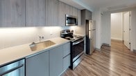 This well-appointed 1-bedroom, 1-bathroom unit offers a thoughtfully designed layout with a consistent modern aesthetic throughout. Hardwood-style flooring flows seamlessly across every room, complementing the clean, neutral color palette of white and cool gray tones.

The open living area is bathed in natural light from large windows, which also frame views of the surrounding cityscape and treetops — a standout feature of this unit. A smart panel near the entryway adds a contemporary, tech-forward touch.

The kitchen is efficiently designed with flat-front cabinetry in a light wood grain finish on top and matte gray below, under-cabinet lighting, a white quartz countertop, a stainless steel single-basin sink, and a full suite of stainless steel appliances including a refrigerator, electric range, microwave, and dishwasher. A white tile backsplash ties the kitchen's modern look together.

The bedroom is generously sized, featuring two large windows that offer sweeping views of the city skyline and surrounding greenery, filling the room with abundant natural light.

The bathroom is clean and minimal, featuring a wall-mounted white vanity, a framed mirror with a floating shelf, and a walk-in tiled shower with a mosaic stone floor — all finished in crisp white tones.