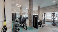 This contemporary fitness center offers a comprehensive workout environment designed to accommodate a wide range of exercise disciplines. The space is anchored by distinctive stone-clad structural columns that add a refined architectural character, complementing the neutral-toned walls and modern finishes throughout.

The flooring features a striking hexagonal tile pattern in teal, beige, and gray, providing both visual interest and a durable, sport-appropriate surface. Overhead, geometric rectangular pendant light fixtures deliver bright, even illumination across the entire floor, enhancing visibility and creating an energetic atmosphere.

The strength training area is outfitted with multiple TKO selectorized machines targeting various muscle groups, alongside a functional trainer cable system and a rack of kettlebells. A floor-to-ceiling mirror wall expands the sense of space and allows users to monitor their form during workouts.

The cardio zone features treadmills, elliptical trainers, upright bikes, and rowing machines, each equipped with individual screens for an engaging workout experience. A large-format tire for functional training rounds out the equipment selection.

Natural light flows in through multiple windows along the perimeter walls, while a glass door provides direct access to the exterior. A small service station with storage and sanitation supplies is conveniently located near the entrance, ensuring a clean and well-maintained environment.