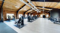Located at 10304 Oxford Hill Dr in Saint Louis, MO, this fitness facility offers an expansive, single-floor workout space distinguished by its unique rustic-modern architectural character. The vaulted tongue-and-groove wood ceiling soars overhead, supported by bold black steel arch beams that run the length of the building, creating a dramatic and inviting atmosphere. Exposed silver ductwork traces the roofline, complementing the industrial aesthetic while natural light filters in through multiple windows along the walls, providing views of the surrounding outdoor greenery.

The generous floor plan is thoughtfully divided into distinct training zones. A dedicated strength training area features rubber flooring and houses a comprehensive array of plate-loaded machines, free weights, dumbbell racks, cable systems, and battle ropes. The cardio zone is equipped with multiple treadmills, ellipticals, and stationary bikes, all positioned on light hardwood-style flooring. A separate functional training area includes rowing machines, spin bikes, and open floor space for dynamic workouts. Large mirrors along one wall enhance the sense of space. The facility's combination of warm wood tones, dark steel accents, and professional equipment creates a welcoming yet high-performance training environment.