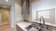 This single-floor unit is designed with a clean, contemporary aesthetic that emphasizes functionality and comfort throughout. The open-concept living area is anchored by light hardwood-style flooring that extends from the kitchen pass-through into the main living space, filling the room with a warm, airy feel. A wall-mounted air conditioning unit and recessed lighting add to the practicality of the space.

The kitchen is well-appointed with flat-front cabinetry in a warm wood-grain finish, white quartz-style countertops, a stainless steel sink with a gooseneck faucet, and a full suite of stainless steel appliances including a range, over-the-range microwave, and refrigerator. A tiled backsplash and a pass-through opening connect the kitchen to the living area, creating a cohesive flow.

The bedrooms are carpeted for comfort and feature neutral walls, standard closets with built-in rods, and windows fitted with horizontal blinds that allow natural light while maintaining privacy. Two bathrooms serve the unit: one features a walk-in shower with white tile surround and a wood-toned vanity, while the second offers a bathtub and similarly neutral finishes. Overall, the space presents a move-in-ready, modern environment with consistent finishes throughout.