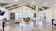 Located at 20 Pine Hall Dr in Clayton, NC, this generously sized clubhouse delivers an impressive blend of architectural character and functional amenity spaces across its expansive single-story footprint. The grand main living area commands immediate attention with its soaring vaulted ceilings accented by green-painted exposed beams and supported by stately white columns. Polished concrete floors reflect abundant natural light pouring in through a dramatic wall of windows, creating an open, airy atmosphere ideal for gathering.

The space flows into a well-appointed kitchen featuring granite countertops, dark cabinetry, stainless steel appliances, a shiplap accent wall, and a large island with bar seating beneath pendant lighting. A separate dining area offers an intimate setting with wainscoting details and colorful artwork. A secondary lounge area provides a more relaxed setting with rich leather seating, crown molding, and wall-mounted media.

An additional room functions as a bright, windowed workspace or meeting room with a large-format roll-up door. A covered outdoor patio with a coffered ceiling and ceiling fan transitions seamlessly to a resort-style pool deck with stone-accented columns and ample lounge seating surrounded by mature tree views.