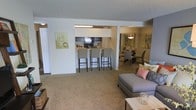 This single-floor residence is designed with an open-concept floor plan that seamlessly integrates the living, dining, and kitchen areas into one cohesive space. The living area features plush carpeting in a warm neutral tone, providing a comfortable foundation for the seating arrangement, which includes a contemporary gray sofa accented with decorative pillows and a light-toned coffee table.

The kitchen is positioned centrally and features a breakfast bar with bar-height seating for three, making it both functional and social. Dark appliances, including a microwave and refrigerator, contrast with the lighter cabinetry, while under-cabinet lighting illuminates the workspace. The pass-through design of the kitchen enhances the sense of openness throughout the unit.

Adjacent to the kitchen, the dining area is defined by an elegant chandelier overhead and accommodates a full dining set, making it well-suited for entertaining. Artwork adorns the walls throughout, adding visual interest against the neutral palette. The textured ceiling and consistent carpeting unify the space, creating a cohesive and inviting atmosphere throughout.