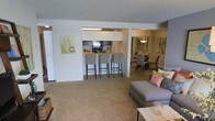 This single-floor residence is designed with an open-concept floor plan that seamlessly integrates the living, dining, and kitchen areas into one cohesive space. The living area features plush carpeting in a warm neutral tone, providing a comfortable foundation for the seating arrangement, which includes a contemporary gray sofa accented with decorative pillows and a light-toned coffee table.

The kitchen is positioned centrally and features a breakfast bar with bar-height seating for three, making it both functional and social. Dark appliances, including a microwave and refrigerator, contrast with the lighter cabinetry, while under-cabinet lighting illuminates the workspace. The pass-through design of the kitchen enhances the sense of openness throughout the unit.

Adjacent to the kitchen, the dining area is defined by an elegant chandelier overhead and accommodates a full dining set, making it well-suited for entertaining. Artwork adorns the walls throughout, adding visual interest against the neutral palette. The textured ceiling and consistent carpeting unify the space, creating a cohesive and inviting atmosphere throughout.