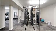 This state-of-the-art fitness center, spanning 930 square feet, is a comprehensive exercise facility designed to cater to various workout needs. The space is thoughtfully divided into distinct zones, each dedicated to specific types of exercise. Upon entry, visitors are greeted by an array of cardio equipment, including treadmills, ellipticals, and stationary bikes, positioned to maximize space efficiency.

The strength training area features a multi-functional cable machine, allowing for a wide range of exercises. Free weights, including dumbbells and barbells, are neatly organized along the walls. A separate section houses weight machines targeting different muscle groups. The center also includes a functional training area with equipment such as stability balls and resistance bands.

The facility boasts modern aesthetics with a neutral color palette, predominantly featuring whites and grays. The flooring consists of durable, low-maintenance carpet tiles in a complementary gray tone. Bright, recessed lighting fixtures ensure optimal visibility throughout the space. Large windows flood the area with natural light and offer views of the outdoors, creating an open and inviting atmosphere.

Wall-mounted televisions and informational signage provide entertainment and guidance for users. The layout promotes easy navigation and maintains clear pathways between equipment. Overall, this fitness center combines functionality with contemporary design, offering a comprehensive workout experience within a compact, well-organized space.