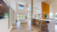 This single-floor community clubhouse is defined by its striking architectural design, centered around dramatically soaring ceilings that create an airy, open atmosphere throughout the space. Towering columns rise to meet the upper reaches of the room, while an array of arched and floor-to-ceiling windows floods the interior with abundant natural light, offering views of lush greenery outside.

The open-plan layout seamlessly connects several functional zones. A curved white reception desk anchors one end of the space, offering a sleek and welcoming point of entry. A central bar-height communal table with warm wood barstools serves as a social hub, illuminated by a series of minimalist pendant lights. A bold amber-orange accent wall provides a striking focal point, housing a wall-mounted flat-screen television.

The lounge area is furnished with plush gold velvet armchairs and contemporary seating arrangements accompanied by marble-based side tables and vertical linear wall sconces. Light wood-look flooring runs throughout, complementing the warm, neutral color palette. Curated artwork and indoor greenery add finishing touches, contributing to the space's inviting and sophisticated ambiance.