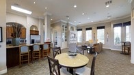 This well-appointed single-floor space offers an open-concept layout that seamlessly connects a kitchen, dining area, and lounge, creating a warm and inviting atmosphere. The interior is defined by refined architectural details, including decorative columns with crown molding, arched built-in niches, and recessed lighting set into smooth white ceilings that enhance the sense of openness.

The kitchen area is equipped with a stainless steel refrigerator and features a raised bar counter with cushioned bar seating, providing a casual spot for gathering. Adjacent to the kitchen, a round dining table surrounded by dark wood chairs sits on a textured tile floor, offering an ideal setting for shared meals or conversation.

The lounge area is furnished with comfortable patterned armchairs and a plush sofa arranged around side tables with warm accent lamps, creating a relaxed and social seating environment. Built-in cabinetry and glass-door display cases add functional elegance, while a television is mounted above a central built-in unit serving as a subtle focal point.

Large windows dressed with dark roller shades and white blinds flood the space with natural light, offering views of lush greenery outside. The neutral color palette of warm beiges and whites, combined with the varied textures of tile flooring and upholstered furnishings, gives the space a sophisticated yet approachable character throughout.
