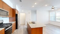 This single-floor space is designed with a clean, modern aesthetic, combining functional layouts with contemporary finishes. The kitchen serves as a central highlight, featuring warm-toned wood cabinetry extending floor to ceiling, light granite countertops, and a central island with a built-in stainless steel sink. Stainless steel appliances — including a gas range, microwave, and refrigerator — complete the well-equipped cooking space. Light wood-look flooring flows consistently throughout the unit, contributing to a bright, cohesive atmosphere.

The living area benefits from natural light through multiple windows fitted with horizontal blinds, while a set of built-in open shelves adds a practical yet attractive architectural detail. Ceiling fans are present in the bedroom areas, enhancing comfort alongside the recessed lighting found throughout.

The bedrooms are generously proportioned, with one featuring access to a walk-in closet with wire shelving. The bathroom is finished with large-format marble-style tile surrounding a soaking tub and shower combination, paired with crisp white fixtures for a polished look. Overall, the space presents a fresh, move-in ready environment with thoughtful, consistent detailing.