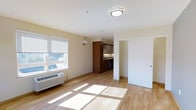 This single-floor space offers a clean, functional layout filled with natural light. The main living area features light hardwood flooring that extends throughout the unit, creating a warm and cohesive aesthetic. A large window fitted with horizontal blinds allows generous sunlight to filter in, casting bright patches across the floor and enhancing the open feel of the room. A wall-mounted HVAC unit provides efficient climate control directly within the living space.

The kitchen is partially open to the living area and showcases rich, warm-toned wood cabinetry paired with gray upper cabinets, offering a pleasing contrast. A stainless steel appliance and a functional countertop with a sink complete the kitchen setup, making it both practical and visually appealing.

A sliding door off the main living area leads to an additional space, providing flexible utility such as a closet or separate room. The walls are finished in a neutral warm white, complementing the natural tones of the flooring and cabinetry. Flush ceiling lighting ensures the interior remains well-lit. Overall, the space presents a tidy, modern, and efficient design that is well-suited for comfortable everyday living.