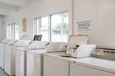 Located in Oakland Park, FL, this single-floor community laundry facility offers a clean, functional, and well-organized space designed to serve multiple users efficiently. The interior is finished with light wood-look flooring and bright white walls, creating a fresh and airy atmosphere throughout.

A row of top-load washing machines lines one wall beneath a series of large windows that invite generous natural light and offer views of the surrounding greenery. On the opposite end, a bank of stacked coin-operated dryers provides ample drying capacity. A spacious central folding table runs along the middle of the room, offering a convenient surface for sorting and folding laundry.

The facility is equipped with ceiling fans and fluorescent lighting, ensuring comfortable conditions year-round. The beamed ceiling adds a subtle architectural character to the otherwise utilitarian space. A seating chair is available for waiting patrons, and a clearly marked exit leads directly to the outdoor parking and grounds area. Safety equipment, including a fire extinguisher, is visibly posted. Informational signage is displayed for user guidance. The open layout and multiple machines make this a practical and accessible shared laundry facility.