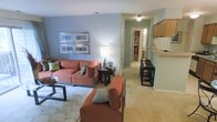 This single-floor residence offers a cohesive, open layout that connects the living area to the kitchen and beyond. The living room is dressed in cool blue-gray walls that create a calm, relaxed atmosphere, complemented by plush neutral carpeting underfoot. The space is furnished with rust-orange upholstered seating, including a sofa and accent chair, both accented with teal throw pillows that echo the wall tones. A patterned area rug defines the seating zone, while a dark wood coffee table and end table with a table lamp add warmth and function. Two framed blue-toned artworks are displayed horizontally on the main wall, contributing a cohesive decorative touch. To the right, the kitchen features a raised breakfast bar with black bar stools, wood-toned upper cabinetry, and light countertops. A hallway extends toward the back of the residence, where a walk-in closet and additional rooms are visible. The open sightlines between the living area, kitchen, and hallway give the space a connected, functional feel throughout.