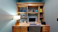 This single-floor office suite offers a thoughtfully designed workspace that balances professional functionality with refined aesthetics. The central meeting area serves as the heart of the suite, featuring a stately dark wood conference table surrounded by teal upholstered chairs. A striking abstract artwork anchors the main wall, while the rich coffered ceiling with warm wood beams and recessed lighting lends an air of sophistication to the room. Wainscoting with raised wood paneling runs along the lower walls, complementing the bold teal paint above and tying together the cohesive design.

Flanking the central area are two private office workstations, each equipped with substantial built-in wood desks and expansive shelving units that provide ample storage and display space. One workstation features a desktop computer setup with a leather task chair, making it fully functional for professional use. Decorative accents — including ceramic vases, small plants, and curated objects — add personality to each workspace.

The suite is accessible via elegant French doors with glass panes that allow natural light to filter in. The flooring throughout features a contemporary multi-toned carpet tile in yellow, green, and grey, adding a modern contrast to the traditional woodwork. This space is well-suited for small meetings, collaborative work, and focused individual tasks.