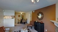 This single-story residence offers an open-concept floor plan that seamlessly connects the living, dining, and kitchen areas into one cohesive and functional space. The living area is anchored by plush carpeting and features a decorative sunburst mirror mounted on a rich, warm-toned accent wall, complemented by a dark wood console cabinet and a patterned accent chair, creating a stylish focal point. A multi-bulb ceiling light fixture provides overhead illumination, while an additional ceiling fan light serves the adjacent dining area.

The dining space accommodates a round dark wood table surrounded by upholstered chairs, making it suitable for everyday dining and casual entertaining. Framed artwork adds a decorative touch to the neutral-toned walls. A glimpse into the kitchen reveals a bar-style counter that opens toward the dining area, offering a convenient pass-through for meals and conversation.

A doorway visible in the background leads to additional rooms, suggesting a practical and well-organized layout. The textured ceiling is consistent throughout, and the overall ambiance is warm, inviting, and thoughtfully furnished with coordinated earth-toned décor.