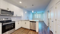 This single-floor unit offers a thoughtfully designed open-concept layout that connects the kitchen and living area into one cohesive space. The kitchen is outfitted with white shaker-style cabinetry, granite countertops, a peninsula with a built-in stainless steel dishwasher and undermount sink, pendant lighting, and a full suite of stainless steel appliances including a range and over-the-range microwave. The living area features warm hardwood-style flooring, a ceiling fan, and windows fitted with horizontal blinds that allow for controlled natural light. A dedicated closet in the main hall conceals a stacked washer and dryer unit alongside additional storage, complemented by a separate double-door closet nearby. The bedroom is carpeted for comfort, includes a ceiling fan, and receives natural light through a window with blinds. The bathroom presents a white vanity with granite countertops, a framed mirror, and hardwood-style flooring, with direct access to a walk-in closet featuring built-in shelving and hanging rods. Clean white walls and modern hardware carry throughout, giving the unit a consistent, contemporary aesthetic.