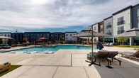 Located at 101–137 Indian Creek Rd NW in Huntsville, Alabama, this outdoor amenity space is the central gathering hub of a contemporary multi-story residential community. The architecture of the surrounding buildings blends dark navy panel siding, white horizontal clapboard, and warm brick accents, creating a visually dynamic backdrop for the outdoor environment.

At the heart of the space is a generously sized, resort-style swimming pool with clear blue water and a wide surrounding deck finished in large-format concrete pavers. The pool deck is furnished with lounge chairs and large cantilever umbrellas, offering ample space for relaxation under the open Alabama sky.

Adjacent to the pool, a dedicated outdoor living area features a built-in brick fireplace with firewood storage, a stainless steel built-in grill station, and comfortable wicker seating arranged on a patterned paver patio. Overhead string lights are strung between poles, providing a welcoming ambiance during evening hours.

A secondary patio courtyard on the opposite side of the complex mirrors this design with additional outdoor seating, dining tables, and grilling stations. The entire pool area is enclosed by a black metal security fence with a gated entry, ensuring controlled access. Landscaping with planters and greenery softens the hardscape throughout.