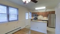 This two-floor space offers a thoughtfully designed layout that balances functionality and comfort. The kitchen is a standout feature, showcasing warm wood cabinetry, light granite countertops, and a tile backsplash that adds texture and visual interest. Stainless steel appliances, including a gas range and built-in microwave, lend a modern, polished feel to the space. A peninsula counter provides additional workspace and serves as a natural dividing point between the kitchen and the adjacent dining or living area. The open layout allows natural light to flow freely through multiple windows fitted with horizontal blinds, creating a bright and welcoming atmosphere. Light hardwood flooring runs throughout the main level, adding warmth and continuity to the space. Crown molding along the ceiling provides a refined architectural detail, while a ceiling fan with a light fixture enhances air circulation and comfort. A baseboard heating system is visible along the walls, ensuring warmth throughout cooler months. A door with a window panel offers access to the exterior, further connecting the interior to the outside. The two-floor configuration offers additional living space beyond the main level, making this a versatile and well-rounded property.