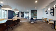 Located at 408 Northampton Rd in Amherst, Massachusetts, this single-floor amenity space is thoughtfully designed to support both collaborative and focused work. The open-plan layout features patterned carpet tile flooring and a clean, contemporary aesthetic defined by a neutral palette accented with deep navy blue walls and bold artwork.

The main area offers multiple seating configurations, including round white pedestal tables paired with modern navy blue chairs, providing flexible gathering spots for small groups. Oversized white dome pendant lights hang above, complementing the recessed ceiling lighting to create a bright, welcoming atmosphere. A large workstation counter with executive-style seating runs along one side of the space, equipped with a multifunction printer, making it suitable for professional tasks.

Natural light filters in through generous windows, adding warmth to the interior. Glass-paneled doors lead to enclosed private rooms, ideal for focused meetings or quiet work sessions. A separate lounge nook features bistro-style black tables with natural wood cane-back chairs, illuminated by decorative wall sconces and adorned with a series of framed abstract artwork in blue and white tones, lending an artistic, refined touch to the space. Overall, this amenity space balances functionality and design to serve a variety of professional and social needs.