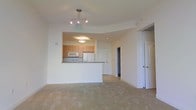 This single-floor space showcases an open-concept design that seamlessly connects the living area to the kitchen. The living area features neutral-toned carpeting and smooth white walls, creating a clean and versatile backdrop. A modern multi-bulb track light fixture is suspended from the ceiling, providing ample illumination throughout the main living space.

The kitchen is defined by a raised peninsula with a granite-style countertop that serves as both a functional workspace and a visual divider between the cooking and living areas. Light wood cabinetry lines the kitchen walls, complemented by a built-in microwave and a white refrigerator. A dedicated overhead light fixture within the kitchen ensures well-lit food preparation. The raised ceiling detail above the kitchen adds architectural interest to the space.

A hallway visible beyond the kitchen leads to additional rooms, with a white paneled door on the right side of the main space providing access to a separate room. The overall ambiance is bright, airy, and move-in ready, with a neutral palette that accommodates a variety of interior styles.