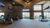 The Fitness Center Cardio room in Conroe, TX is a well-appointed, single-floor facility designed to support an effective cardiovascular workout experience. The space is immediately distinguished by its dramatically high ceilings, which lend an open, airy quality and contribute to a comfortable exercise environment. Large floor-to-ceiling windows along the exterior walls flood the interior with generous natural light while offering views of the surrounding community and landscaping. Clerestory windows positioned near the upper portion of the walls further enhance brightness throughout the day.

The flooring consists of durable, patterned carpet tile suited for high-traffic fitness use. Cardio equipment is thoughtfully arranged throughout the space and includes treadmills, elliptical machines, stationary bikes, and additional cardio-focused machines. Multiple wall-mounted flat-screen televisions are positioned at comfortable viewing heights across the room, allowing users to stay entertained during workouts. Full-length mirrors along one wall visually expand the space and support proper form during exercise.

The neutral warm-toned walls are complemented by gray-accented louvered interior doors, providing access to utility areas. A hand sanitizer dispenser and thermostat are conveniently located on the wall. The entrance features glass doors clearly marked as the Fitness Center, ensuring easy access and a welcoming, professional atmosphere.