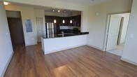 This single-floor space showcases a thoughtfully designed open-concept layout that seamlessly integrates the kitchen and living areas. The kitchen is a standout feature, highlighted by rich dark cabinetry that contrasts elegantly with a bold black granite countertop. A peninsula with a white base extends into the living space, offering additional prep space and a natural gathering point. Stainless steel appliances, including a full-sized refrigerator, complement the modern aesthetic, while a trio of pendant lights suspended from a track rail above the peninsula adds focused task lighting and a decorative touch. The kitchen also benefits from under-cabinet lighting and additional recessed track fixtures, ensuring the workspace is well-illuminated. Warm-toned hardwood flooring runs throughout the open living area, adding natural texture and continuity to the space. The neutral, light-colored walls enhance the bright and airy atmosphere, while standard-height ceilings maintain a comfortable, approachable scale. A paneled interior door is visible, suggesting a functional layout with defined zones for additional rooms beyond the open living and kitchen area.
