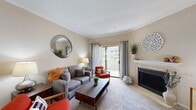 This single-floor residence offers a thoughtfully designed layout that balances comfort and functionality. The living room serves as the heart of the home, featuring warm neutral tones, crown molding, and plush carpeting. A tile-surround fireplace anchors one wall, while a sliding glass door with grayish curtains opens to a private balcony, flooding the space with natural light and providing outdoor views. The open-concept layout connects the living area to the kitchen, which is outfitted with rich warm wood cabinetry, granite-style countertops, white appliances including a range, microwave, refrigerator, and dishwasher, and durable hardwood-style flooring. A breakfast bar provides a casual dining option between the kitchen and living areas. The bedroom is a serene retreat with soft carpeting, a ceiling fan, ample closet space, and a door with shuttered windows that allows natural light in. The bathroom features a granite countertop vanity with an undermount sink, a tub-shower combination with marble-style tile surround, and a large mirror. Throughout the residence, warm beige walls and crown molding contribute to a cohesive and inviting ambiance.