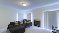 This single-floor space presents a well-appointed living room that balances comfort and functionality. The room is finished with light-toned carpeting that adds warmth underfoot, while white walls and a textured ceiling contribute to a clean, bright aesthetic. A centrally positioned flush-mount ceiling light provides consistent overhead illumination throughout the space.

The living area's standout feature is a tile-surround fireplace with a sleek black insert, serving as a natural focal point along one wall. Large vertical-blind-covered windows line the walls, allowing for adjustable natural light and privacy control. The generous layout comfortably accommodates a large sectional sofa with decorative accent pillows, complemented by a compact side table for added functionality.

The overall ambiance is neutral and move-in ready, offering a versatile canvas suitable for a variety of interior design preferences. The open arrangement of the living space encourages easy furniture placement and a relaxed, welcoming atmosphere ideal for everyday living and entertaining.