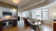 This single-floor urban residence offers a well-considered layout that balances open communal spaces with private retreats. The living area is a highlight of the unit, defined by expansive floor-to-ceiling windows that frame sweeping city skyline views and flood the space with natural light. Exposed cylindrical ductwork runs along the ceiling, lending an industrial-contemporary character that distinguishes the architectural design. Light hardwood flooring flows throughout, unifying the living, dining, and kitchen areas into a cohesive open-plan arrangement. A dedicated dining nook with access to an outdoor balcony extends the living space further.

The kitchen is outfitted with stainless steel appliances—including a range, microwave, and dishwasher—paired with two-tone cabinetry featuring white upper cabinets and dark lower cabinets, set against clean light countertops.

The primary bedroom benefits from corner windows with urban views, while a second bedroom offers a quieter retreat with its own city outlook. The bathroom is clean and functional, finished with light wood-toned flooring, a single-sink vanity, and a shower enclosure.

A hallway connects the private areas and features a stacked in-unit washer and dryer, adding everyday convenience. Throughout, the space maintains a bright, modern ambiance enhanced by thoughtful natural light and urban vistas.