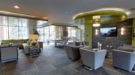 This contemporary community lounge is a thoughtfully designed gathering space that blends modern aesthetics with functional amenities. The open-plan layout is anchored by a striking curved ceiling detail finished in bold chartreuse green, which adds a vibrant pop of color and architectural interest to the room. Recessed lighting, pendant fixtures, and an elegant crystal chandelier work together to create a warm, inviting ambiance throughout.

The space is divided into distinct activity zones. A central billiards table serves as a social focal point, while a dedicated dining or meeting area in the background offers a more structured setting for group gatherings. Comfortable lounge seating, including modern upholstered armchairs in soft grey tones, is arranged to encourage conversation and relaxation.

A striking stacked stone accent wall adds a natural, textural contrast to the otherwise sleek interior, complemented by wall-mounted artwork and decorative sconces. Floor-to-ceiling windows along the perimeter flood the space with abundant natural light and offer views to the outdoors, creating a seamless connection between the interior and exterior environments. Patterned carpeting grounds the space and adds depth to the overall design. Together, these elements create a sophisticated, multi-purpose lounge suited for both casual and social use.