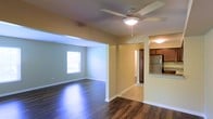 This single-story space features an open-concept layout that connects the main living area to the kitchen, creating a cohesive and functional flow throughout. The living room is finished with dark hardwood-style flooring that adds warmth and contrast to the neutral, tan-toned walls. A ceiling fan with integrated lighting is centrally mounted overhead, providing both airflow and ambient illumination. A window along the far wall allows natural light to fill the space.

Adjacent to the living area, the kitchen is outfitted with dark wood cabinetry, a stainless steel refrigerator, and granite-style countertops. A pass-through or raised counter connects the kitchen visually to the main living space, enhancing the open feel. The kitchen features light tile flooring, offering a clean contrast to the darker flooring of the living area.

A hallway visible from the main living space leads to additional areas of the unit, suggesting a well-organized floor plan. White baseboards throughout complement the neutral wall tones, adding a clean, finished look. Overall, the space presents a practical and inviting atmosphere suited for everyday living.