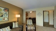 This single-floor space showcases an open-concept layout that seamlessly connects the living room, dining area, and kitchen. The living room features warm, neutral-toned walls with a contrasting olive-green accent wall, creating a cozy and inviting atmosphere. Plush carpeted flooring runs throughout, adding comfort underfoot. A floor lamp provides warm ambient lighting, complementing the natural color palette of the furnishings, which include a grey sofa and a patterned accent chair with an ottoman.

An open archway leads into the dining area, where a ceiling fan with integrated lighting adds both functionality and airflow. A dark-wood dining table with matching chairs provides a dedicated space for meals. The kitchen, visible beyond the dining area, features wooden cabinetry and a functional layout suited for everyday cooking needs.

A decorative wall clock adds a subtle design touch to the living space, while the open flow between rooms enhances the sense of spaciousness. The overall ambiance is practical and comfortable, with a cohesive warm-toned design scheme that ties each area of the home together.