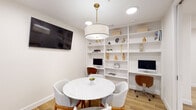 This thoughtfully designed co-working space blends modern aesthetics with practical functionality. The room is anchored by a striking round marble-top table surrounded by four upholstered mid-century modern chairs, ideal for small group meetings or collaborative work sessions. A statement drum pendant light with a brushed brass fixture hangs overhead, complementing the recessed lighting that keeps the space bright and welcoming.

Spanning an entire wall, floor-to-ceiling built-in white shelving provides ample storage and display space, with decorative objects and curated accessories adding warmth and personality. Integrated into the shelving unit are two dedicated workstations, each equipped with a desktop computer and an ergonomic mid-century style chair, offering residents private spots for focused individual work. Wi-Fi connectivity is clearly designated within the space.

A wall-mounted flat-screen television provides an additional screen resource for presentations or media. The flooring throughout features wide-plank wood-look tile in a warm honey tone, enhancing the room's inviting ambiance. Full-height glass partition walls with sleek black framing offer a sense of openness and visual connectivity to the surrounding corridor and adjacent lounge areas, maintaining privacy while preserving an airy, transparent feel throughout.