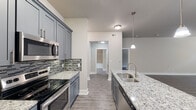 This well-designed single-floor residence offers a cohesive modern aesthetic throughout, anchored by a standout kitchen that serves as the heart of the home. The kitchen is appointed with sleek gray shaker-style cabinetry, granite countertops, a mosaic tile backsplash, and a full suite of stainless steel appliances including an electric range, built-in microwave, and dishwasher. A central island with a undermount sink and pendant lighting above provides both additional prep space and a natural gathering point that opens toward the living area. Light gray wood-look flooring runs consistently throughout, lending a clean and unified feel to the entire space. The bedrooms are generously proportioned, each featuring ceiling fans, horizontal-slat window blinds that allow natural light while maintaining privacy, and neutral wall tones that create a calm, versatile atmosphere. Storage is well-addressed, with a walk-in closet offering multiple hanging rods and shelving. The primary bathroom impresses with a dual-sink granite vanity, gray cabinetry with brushed nickel hardware, a large framed mirror, and a separate tiled shower. A secondary bathroom mirrors the refined finishes, featuring a granite vanity and a tiled tub-shower combination set within an elegant arched alcove. Overall, the space presents a polished, move-in-ready interior with consistent, quality finishes throughout.