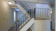 This two-story space is defined by a prominent central staircase that serves as both a functional and visual focal point. The staircase features carpet-lined treads and is framed by clean black metal railings with a straightforward vertical baluster design, lending a practical yet modern industrial character to the interior. The stairs are arranged in a switchback configuration, efficiently connecting the lower and upper levels while maintaining an open, airy feel between floors.

The surrounding walls are finished in a crisp white, contributing to a bright and clean atmosphere throughout the space. On the lower level, dark hardwood-style flooring contrasts with the carpeted stair treads, adding visual interest and definition between areas. Natural light filters in through windows visible on the lower floor, enhancing the openness of the layout.

The upper level features a carpeted landing area enclosed by matching black metal railings, providing a sense of continuity in design between floors. The open railing system allows sightlines across both levels, reinforcing the spacious, connected feel of the interior. The textured ceiling and overall neutral color palette create a versatile backdrop suitable for a variety of uses. Overall, the space presents a functional, well-lit two-level layout with consistent design details that tie both floors cohesively together.