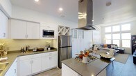 This single-floor space showcases a thoughtfully designed open-concept layout where the kitchen serves as a central hub. The kitchen is outfitted with crisp white cabinetry, grey countertops, and stainless steel appliances, including a full-size refrigerator and a built-in microwave. A standout feature is the large central island equipped with a gas cooktop and a sleek stainless steel range hood suspended above, serving both functional and aesthetic purposes. The cabinetry includes a distinctive X-pattern wine rack, adding a refined touch to the storage options. A subway tile backsplash runs along the perimeter walls, complementing the clean, contemporary palette. Frosted glass upper cabinets near the transition zone add a subtle decorative element while maintaining a light, airy feel throughout. Recessed lighting illuminates the space efficiently, and a window brings in natural light. The kitchen opens fluidly into the living area, where a comfortable seating arrangement is visible, reinforcing the cohesive and connected nature of the overall layout.