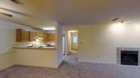 This single-floor residence offers a practical and comfortable layout designed for easy living. The open-concept design connects the kitchen and living areas seamlessly, creating a sense of spaciousness throughout. The kitchen features warm, medium-toned wood cabinetry, a white refrigerator, and a built-in range with an overhead ventilation hood. A pass-through counter with a breakfast bar opens directly into the living space, making the kitchen feel accessible and social. Flush-mounted fluorescent lighting illuminates the kitchen work areas effectively. The living area is finished with soft, neutral-toned wall-to-wall carpeting that extends through the hallway, adding warmth and comfort underfoot. Walls throughout are painted in a clean, light neutral tone that enhances the brightness of the space. A hallway leads toward the entryway, where a windowed door allows natural light to filter into the interior, providing a welcoming transition point. The overall ambiance of the space is cozy and functional, with a straightforward floor plan that maximizes usability across all areas of the home.