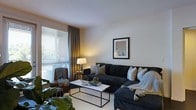 This single-floor space features a thoughtfully designed living area that balances comfort and contemporary style. The room centers around a large, dark navy sectional sofa adorned with decorative pillows in neutral and patterned tones, complemented by a draped throw for added warmth. A sleek marble-top coffee table with a minimalist black metal frame serves as a functional focal point, styled with books and geometric decorative accents.

Natural light filters through wide horizontal blinds on a prominent window, dressed with floor-length charcoal curtains that add depth and elegance. A warm-toned table lamp on a side table enhances the ambient lighting. A classic brown leather armchair with a casual throw sits beside the window, creating a relaxed reading nook.

The walls are kept in soft, neutral tones, providing a clean backdrop for a framed piece of wall art that adds a subtle pop of color. Lush indoor plants near the foreground introduce natural texture and vibrancy to the space. The overall ambiance is warm, inviting, and cohesively styled with modern yet comfortable furnishings.