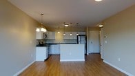 This single-floor residence offers a thoughtfully designed open-concept layout that seamlessly connects the kitchen and living areas. The kitchen is a standout feature, showcasing white cabinetry, dark countertops, and stainless steel appliances including a refrigerator, stove, and built-in microwave. A central island provides additional workspace and serves as a natural dividing point between the kitchen and the open living area. Warm hardwood flooring flows throughout the space, lending a cohesive and inviting atmosphere. Lighting plays a key role in defining the space — an elegant chandelier anchors the dining area, while a series of pendant lights hang above the kitchen, adding both function and visual interest. The ceilings are smooth and well-lit with recessed fixtures that brighten the entire area evenly. Neutral wall tones in a soft warm beige complement the light cabinetry and flooring, creating a balanced and welcoming palette. A doorway visible at the rear of the space leads to additional rooms, suggesting a practical and well-organized floor plan. Overall, the space combines modern finishes with a comfortable, open feel suited for everyday living.