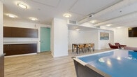 Located at 600 E Superior Ave in Cleveland, Ohio, this single-floor amenity lounge is thoughtfully designed to accommodate socializing, recreation, and casual gatherings. The space is divided into several distinct functional zones, all unified by light wood-look flooring, crisp white walls, and consistent flush-mount lighting throughout.

The lounge area anchors the space with a comfortable sectional sofa, deep burgundy accent chairs, a patterned area rug, and a wall-mounted flat-screen television, creating an inviting setting for relaxed conversation or entertainment. Bold urban-themed artwork adds character to the walls and reinforces the space's connection to its Cleveland surroundings.

Adjacent to the lounge is a dining area featuring a sleek table with tan upholstered chairs, suitable for group meetings or casual meals. The game zone includes an air hockey table, offering a recreational focal point for residents. A kitchenette with dark wood cabinetry, stone-look countertops, and a beverage refrigerator provides convenient amenity support for gatherings.

Windows along the perimeter bring in natural light and frame views of the surrounding downtown Cleveland cityscape. The open, interconnected layout and contemporary furnishings throughout give the space a clean, welcoming, and versatile atmosphere.