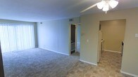 This single-floor residence offers an open-concept layout that connects the main living area to the kitchen and additional rooms. The living space features wall-to-wall carpeting and is painted in a neutral gray-green tone, creating a calm, understated ambiance. A ceiling fan with a light fixture is centrally positioned, providing both airflow and overhead lighting. Vertical blinds on the window allow for adjustable natural light. HVAC vents are visibly integrated into the walls and ceiling throughout.

A partial wall divides the living area from the kitchen, which transitions to tile flooring in a warm earth-tone pattern. The kitchen is equipped with standard appliances, including a refrigerator and range, and features light-colored cabinetry. The layout allows a natural flow between the kitchen and living spaces.

A doorway visible from the main room leads to what appears to be a bedroom, featuring a door with a bottom-mounted vent. White baseboards run along the perimeter of each room, adding a clean contrast to the neutral walls. The overall design is functional and straightforward, suited for practical everyday living.