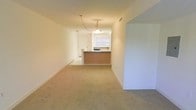 This single-floor space offers a straightforward, functional layout with a seamlessly connected living area and kitchen. The living area is finished with wall-to-wall beige carpeting, which lends a neutral, understated aesthetic throughout. Crisp white walls and white baseboards contribute to a clean, bright atmosphere.

The kitchen is positioned at the far end of the space and is defined by a raised peninsula counter with a warm-toned base, providing a natural visual boundary between the cooking and living areas. The kitchen features dark countertops that contrast with the lighter surroundings, and a window above the sink allows natural light to filter into the space. A pendant light fixture hangs above the peninsula, offering both functional task lighting and a subtle focal point. A doorway adjacent to the kitchen provides access to additional areas of the unit.

The open-plan arrangement between the living and kitchen areas promotes a sense of connectivity and ease of movement. The overall ambiance is calm and neutral, offering a versatile canvas suitable for a variety of interior design approaches.