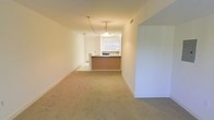 This single-floor space offers a straightforward, functional layout with a seamlessly connected living area and kitchen. The living area is finished with wall-to-wall beige carpeting, which lends a neutral, understated aesthetic throughout. Crisp white walls and white baseboards contribute to a clean, bright atmosphere.

The kitchen is positioned at the far end of the space and is defined by a raised peninsula counter with a warm-toned base, providing a natural visual boundary between the cooking and living areas. The kitchen features dark countertops that contrast with the lighter surroundings, and a window above the sink allows natural light to filter into the space. A pendant light fixture hangs above the peninsula, offering both functional task lighting and a subtle focal point. A doorway adjacent to the kitchen provides access to additional areas of the unit.

The open-plan arrangement between the living and kitchen areas promotes a sense of connectivity and ease of movement. The overall ambiance is calm and neutral, offering a versatile canvas suitable for a variety of interior design approaches.