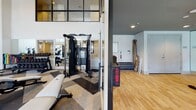 This contemporary fitness center is designed to support a full range of workout routines across two distinct zones. The main strength and cardio area boasts soaring double-height ceilings and expansive floor-to-ceiling windows that flood the space with natural light and offer open views of the surrounding exterior. The patterned carpet tile flooring in a bold chevron design adds a dynamic visual element underfoot, while pendant lighting and wall-mounted TVs contribute to a polished, modern atmosphere. This zone is outfitted with a comprehensive selection of equipment, including ellipticals, stationary bikes, rowing machines, treadmills, a multi-functional cable and strength training rig, free weights with medicine balls, and a Treadwall climbing machine. Full-length mirrors amplify the sense of space throughout. A separate cycling studio is accessed through glass-paneled doors and features smooth hardwood-style flooring, rows of stationary spin bikes, mirrored walls, and a large display screen at the front of the room — ideal for group fitness classes. An additional flex area with hardwood flooring provides space for stretching, mat work, or accessory storage. Together, the zones create a comprehensive, well-lit fitness facility that balances high-energy training with thoughtful design.