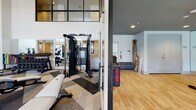 This contemporary fitness center is designed to support a full range of workout routines across two distinct zones. The main strength and cardio area boasts soaring double-height ceilings and expansive floor-to-ceiling windows that flood the space with natural light and offer open views of the surrounding exterior. The patterned carpet tile flooring in a bold chevron design adds a dynamic visual element underfoot, while pendant lighting and wall-mounted TVs contribute to a polished, modern atmosphere. This zone is outfitted with a comprehensive selection of equipment, including ellipticals, stationary bikes, rowing machines, treadmills, a multi-functional cable and strength training rig, free weights with medicine balls, and a Treadwall climbing machine. Full-length mirrors amplify the sense of space throughout. A separate cycling studio is accessed through glass-paneled doors and features smooth hardwood-style flooring, rows of stationary spin bikes, mirrored walls, and a large display screen at the front of the room — ideal for group fitness classes. An additional flex area with hardwood flooring provides space for stretching, mat work, or accessory storage. Together, the zones create a comprehensive, well-lit fitness facility that balances high-energy training with thoughtful design.