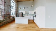 Located at 122 E Main St in Haw River, NC, this single-floor loft showcases a compelling blend of industrial heritage and contemporary design. The space is defined by its dramatic soaring ceilings with exposed ductwork and original wood ceiling planks, lending an authentic mill-building character throughout.

The open living area is lined with a series of large, arched multi-pane windows set within richly textured, weathered exposed brick walls — flooding the interior with natural light and offering scenic outdoor views. Original hardwood floors run throughout much of the space, complementing the raw, historic aesthetic, while polished concrete flooring in the bedroom area adds a modern industrial contrast.

The kitchen is a standout feature, outfitted with crisp white shaker cabinetry, a striking marble-veined quartz island, a subway-style backsplash in soft sage, stainless steel appliances, and stylish pendant lighting. Original structural columns with layers of patinated paint remain as artistic reminders of the building's storied past.

The bathroom is finished with clean, modern marble-look tile in a glass-enclosed walk-in shower. Throughout the space, the interplay of preserved raw materials and thoughtful modern updates creates a one-of-a-kind living environment with unmistakable character and warmth.