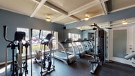 Located at 9010 Markville Dr in Dallas, Texas, this fitness center offers a well-equipped, single-floor exercise facility designed to support a full range of workout routines. The space is finished with cool blue-gray walls and elegant coffered ceilings with built-in lighting, creating a clean and motivating atmosphere throughout.

The cardio zone is lined with multiple treadmills and elliptical machines from SportsArt, positioned along a wall of large windows that invite abundant natural light and provide pleasant outdoor views of the surrounding landscaped grounds. Full-length mirrors along one wall visually expand the space and allow for form checks during exercise.

The strength training area includes a multi-station cable machine and a dedicated free weight section with a tiered dumbbell rack offering a range of weights, accompanied by a flat utility bench. A stability ball is also available for functional fitness exercises. Rubber flooring throughout provides durability, safety, and comfort underfoot.

The entry area features posted exercise facility policies, ensuring users are well-informed before beginning their workouts. Track lighting near the entrance and ample ceiling fixtures ensure consistent illumination across all zones. The overall layout is open and thoughtfully organized, making efficient use of the available floor space while maintaining clear pathways between equipment zones.